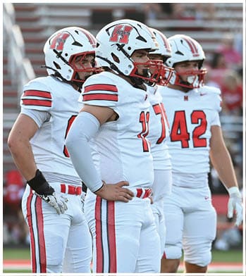 Three people in white football uniforms with red and black stripes stand on a green field during a game. Description generated by AI