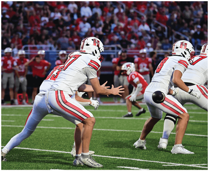 People in white and red football uniforms are positioned on a green field with a crowd in the background. Description generated by AI