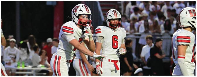Three people in white football uniforms with red and black stripes are on a field, with one person gesturing toward another. Description generated by AI
