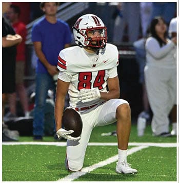 A person wearing a white football uniform with the number 84 and a helmet is holding a football on a field with blurred people in the background. Description generated by AI