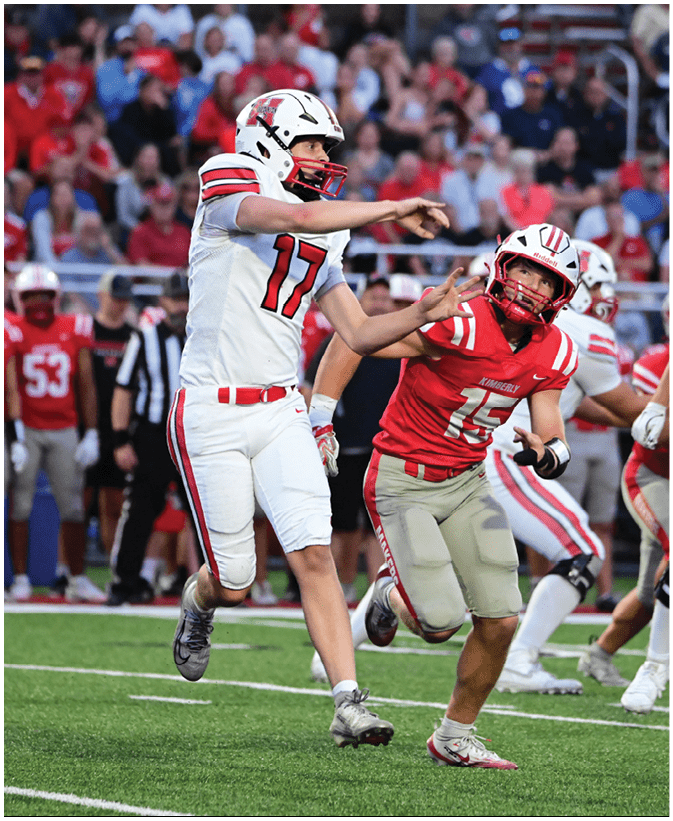 Two people in football uniforms are actively engaged in a play on a field with one person in white throwing a ball and the other in red attempting to block or intercept. Description generated by AI