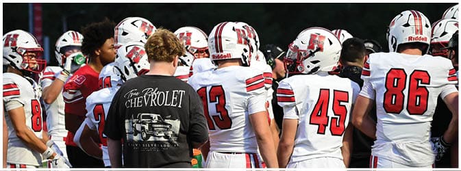 People wearing football uniforms and helmets are standing together on a field with one person in a black Chevrolet shirt among them. Description generated by AI