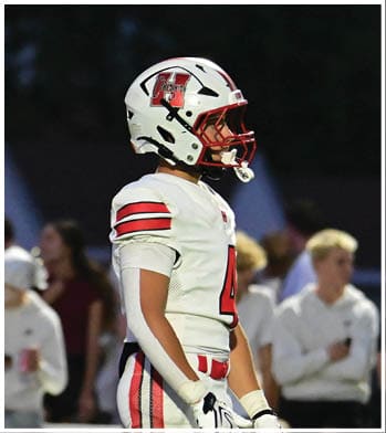 A person in a white football uniform and helmet stands on a field with a crowd of people in casual clothing in the background. Description generated by AI