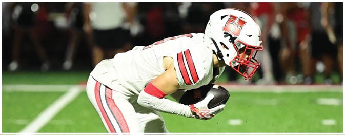 A person in a white football uniform with red stripes is crouched on a green field holding a football, preparing for a play. Description generated by AI