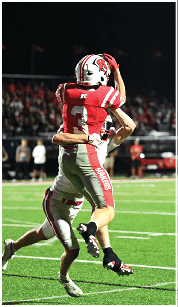 A person wearing a red football uniform with the number 3 and a white helmet is raising one hand to their head on a dark background with blurred spectators. Description generated by AI