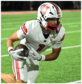 A person wearing a white football uniform with red and black stripes is holding a football on a green field. Description generated by AI