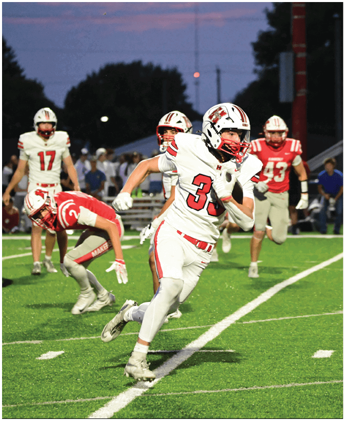 A person wearing a white football uniform with the number 3 is running on a field while several other players in red and white uniforms are behind. Description generated by AI