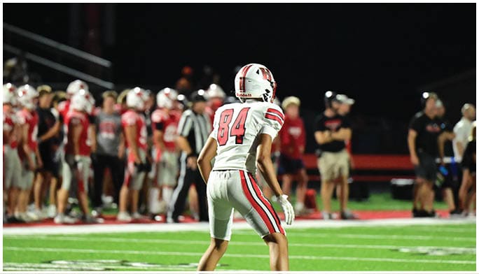 A person wearing a white sports uniform with the number 84 is on a field at night while a group of people stand in the background. Description generated by AI