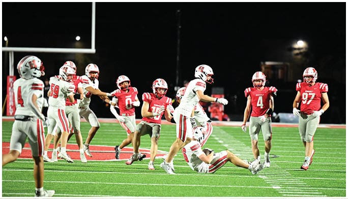 People wearing football uniforms are actively playing on a well-lit field at night, with one person on the ground and others running or standing nearby. Description generated by AI