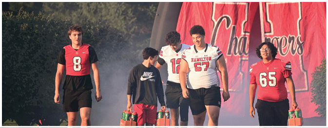 Five people wearing sports jerseys and casual clothing walk together outdoors near a large red banner with white lettering. Description generated by AI
