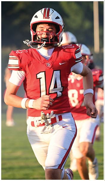 A person wearing a red and white football uniform with the number 14 is running on a grassy field with other players in the background. Description generated by AI