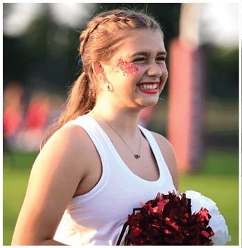 A person with braided hair is smiling and holding red and white pom-poms while wearing a white tank top and face glitter. Description generated by AI