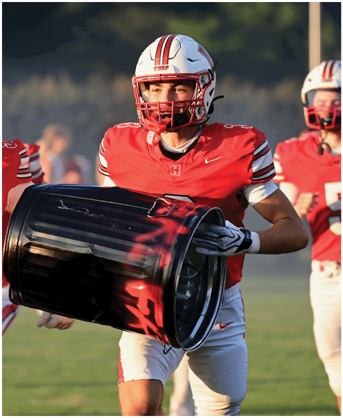 People in red and white football uniforms are running on a field, with one person in the foreground holding a large black drum. Description generated by AI