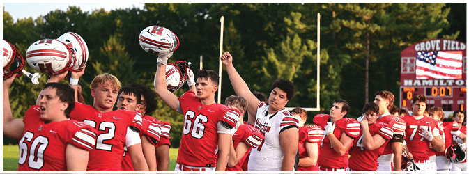 A group of people wearing red and white football uniforms stand in a line outdoors, some holding their helmets up, with a scoreboard and goalposts visible in the background. Description generated by AI