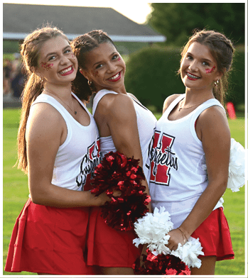 Three adults wearing matching cheerleading outfits with red skirts and white tops hold red and white pom-poms while smiling outdoors on a grassy field. Description generated by AI