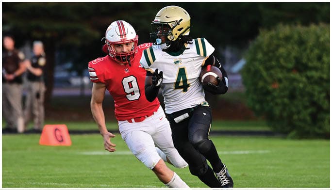 Two people in football uniforms are on a field with one person in a white and green jersey holding a football while the other in a red jersey is closely following. Description generated by AI
