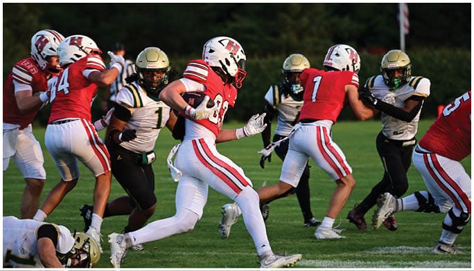 People wearing football uniforms are actively playing on a grassy field with one person holding the ball and others running nearby. Description generated by AI