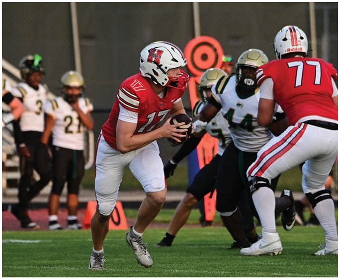 People in football uniforms are actively playing on a grassy field with one person holding the ball and others preparing to block or tackle. Description generated by AI