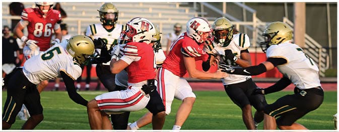 Adults in football uniforms are actively engaged in a game on a field with bleachers in the background. Description generated by AI