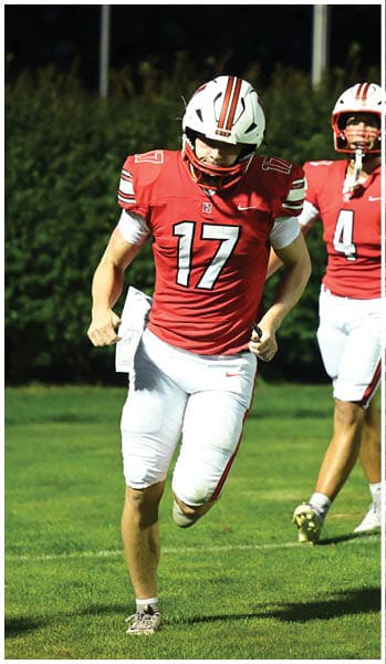 Two people wearing red and white football uniforms and helmets are on a grassy field, with one running toward the camera and the other standing behind. Description generated by AI
