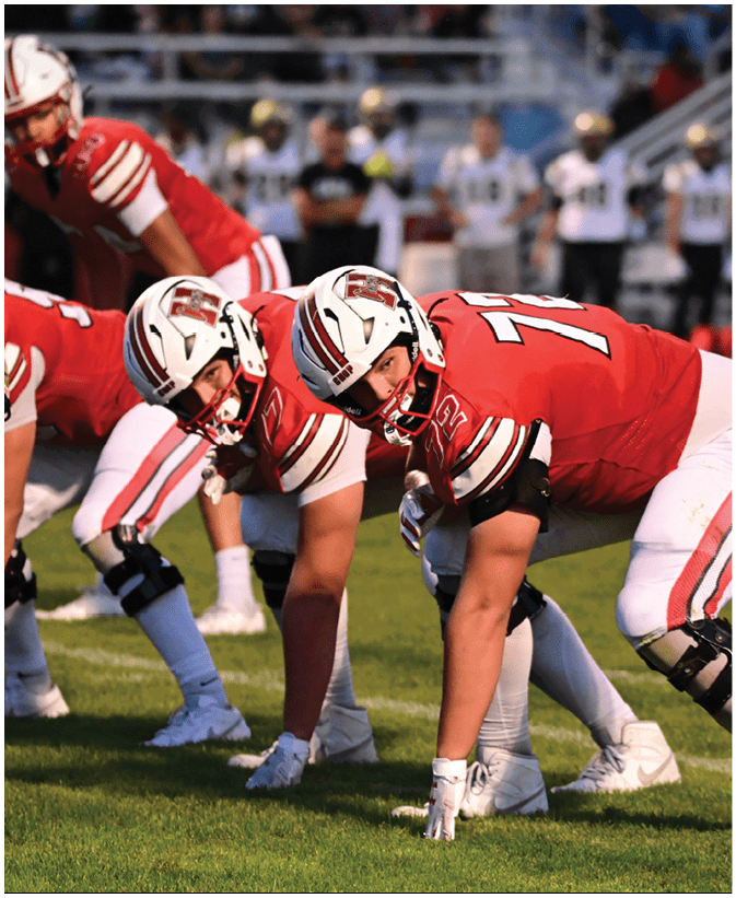 People in red and white football uniforms are lined up on a grassy field preparing for a play during a game. Description generated by AI