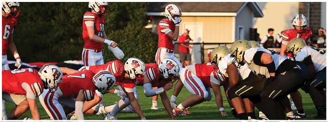 Two opposing American football teams in helmets and uniforms are lined up facing each other on a grassy field preparing for a play. Description generated by AI