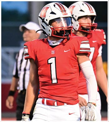 A person wearing a red football uniform with the number one and a white helmet stands on a field with a referee in the background. Description generated by AI