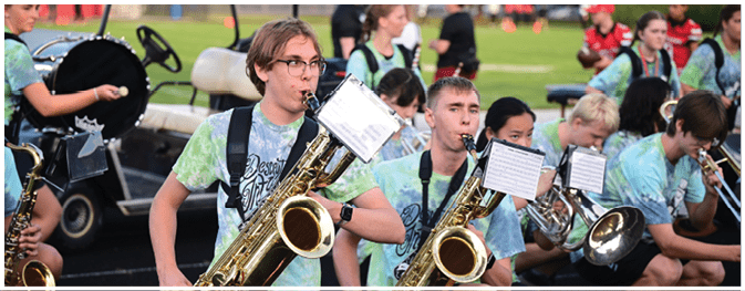 People wearing matching tie-dye shirts are seated outdoors playing large brass saxophones with sheet music attached. Description generated by AI