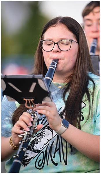 A person with long dark hair and glasses is playing a wind instrument outdoors. Description generated by AI