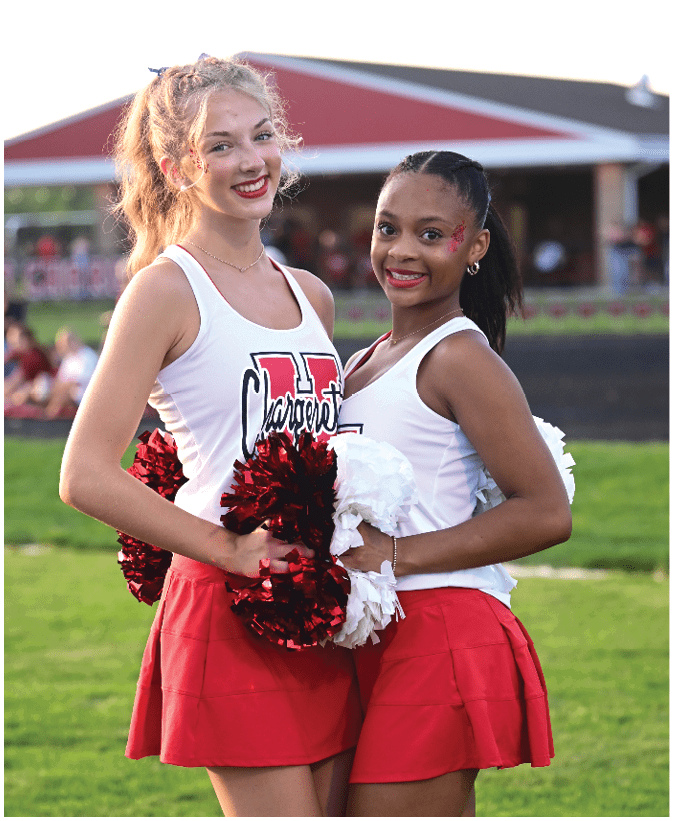Two adults wearing cheerleading uniforms with red and white pom-poms smile while standing outdoors near a building with a red roof. Description generated by AI