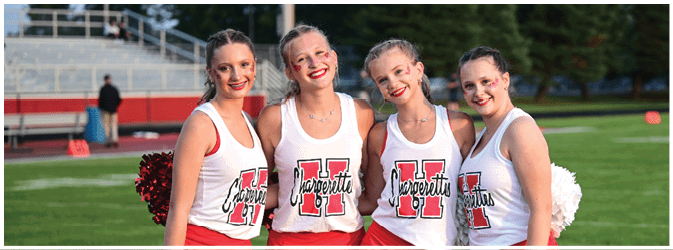 Four adults wearing matching cheerleading outfits with white tops and red skirts stand closely together on a grassy field, smiling and holding pom-poms. Description generated by AI
