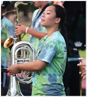 People wearing tie-dye shirts are playing brass and woodwind instruments outdoors, with one person in the foreground holding a euphonium. Description generated by AI