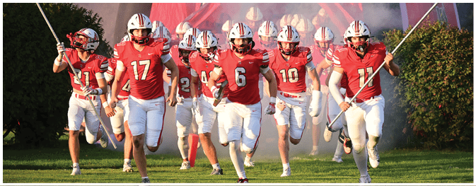 A group of people wearing red sports jerseys and white pants are running onto a grassy field holding flags. Description generated by AI