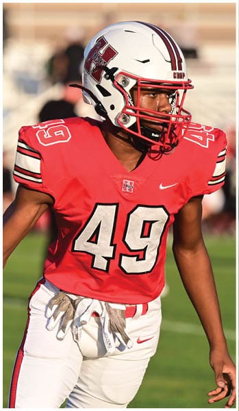 A person wearing a red football jersey with the number 49 and a white helmet is holding a football while standing on a grassy field. Description generated by AI