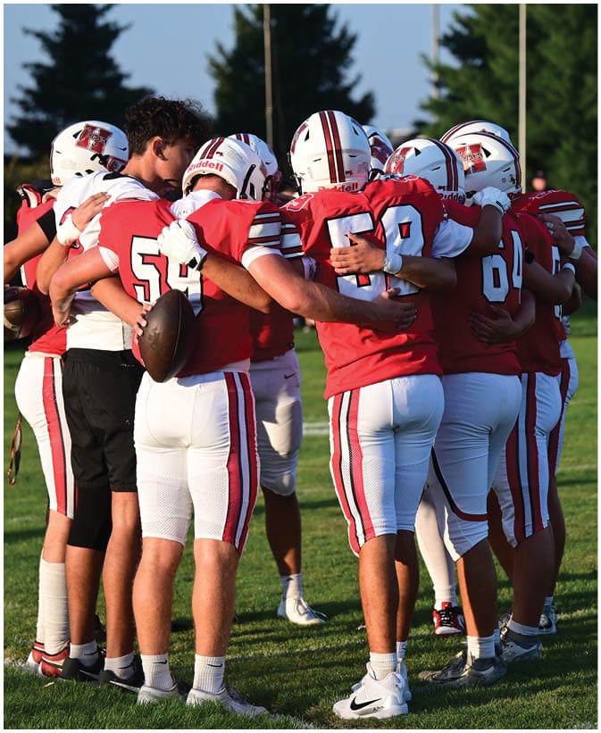 A group of people wearing red and white football uniforms are standing in a close huddle on a grassy field with trees and light poles in the background. Description generated by AI