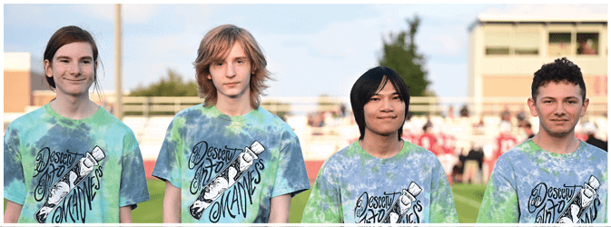 Four people wearing matching tie-dye shirts with a graphic design stand outdoors on a grassy field with a blurred crowd in the background. Description generated by AI