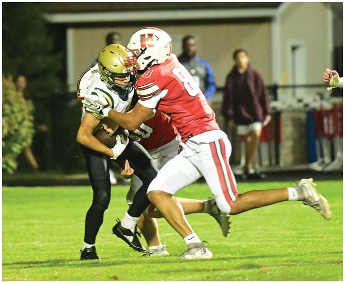 Two people in football uniforms are engaged in a tackle on a grassy field at night with a small building and spectators in the background. Description generated by AI