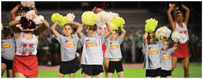 Children wearing gray shirts with “Kids Clinic" printed on them are holding colorful pom-poms and performing a cheer routine on a field. Description generated by AI