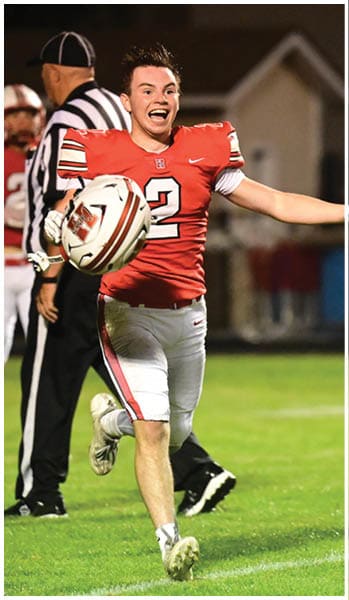 A person wearing a red football jersey and white pants is running on a grassy field with an excited expression while holding a football. Description generated by AI