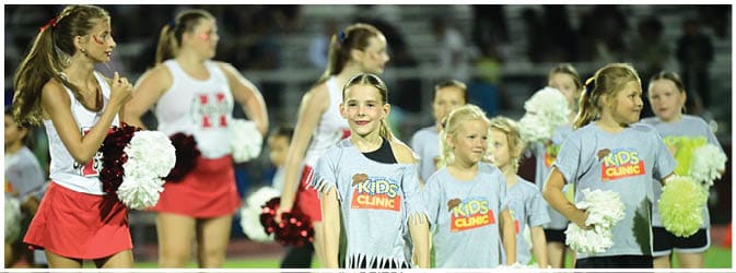 Children wearing cheerleading outfits and t-shirts with “Kids Clinic" printed on them are standing on a sports field holding pom-poms. Description generated by AI