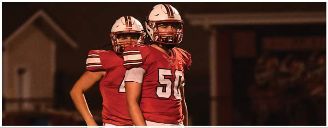 Two people wearing red football uniforms and helmets stand on a field at night. Description generated by AI