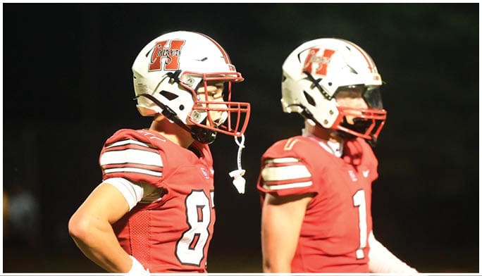 Two people wearing red football uniforms and white helmets stand side by side on a field at night. Description generated by AI