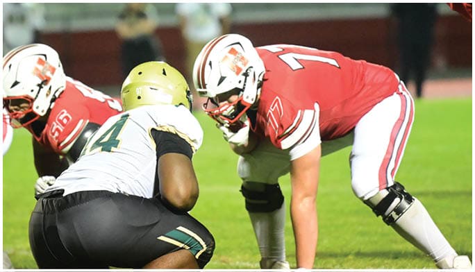 Several football players in red and white uniforms are lined up on the field facing a player in a white and black uniform in a crouched position during a game. Description generated by AI