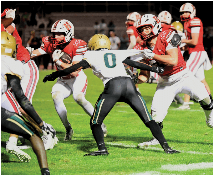 Two people in football uniforms are engaged in a play on a grassy field with one player in red and white grabbing the jersey of another player in black and white. Description generated by AI