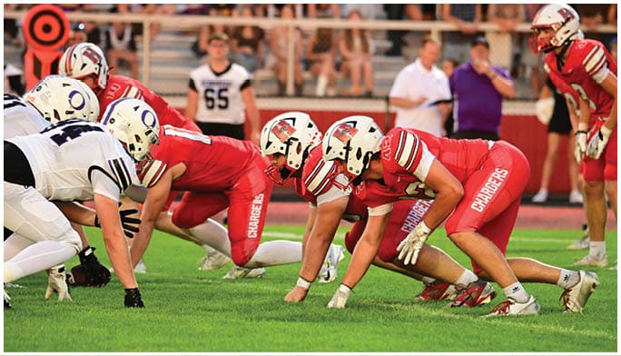 Three athletes in red football uniforms and helmets are crouched in a ready position on a grassy field during a game. Description generated by AI