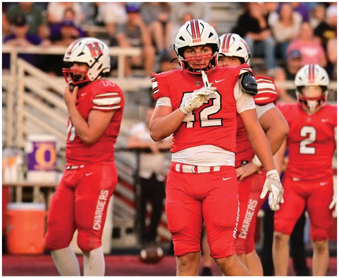 People in red football uniforms and helmets stand on a field with a blurred crowd in the background. Description generated by AI