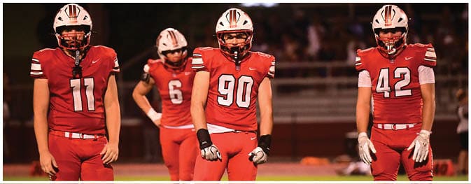 Four people wearing red football uniforms with helmets stand on a field, with three in the foreground and one slightly behind. Description generated by AI