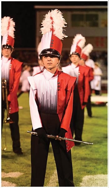Person wearing a red and white marching band uniform with a tall hat adorned with white feathers standing outdoors. Description generated by AI