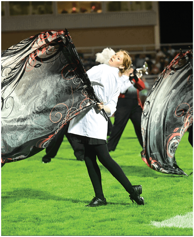 A person in a white jacket and black leggings is performing with a large decorative flag on a grassy field, with other performers and a crowd visible in the background. Description generated by AI