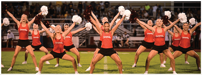 A group of adults wearing matching red tops and black shorts perform a synchronized cheerleading routine on a grassy field while holding pom-poms. Description generated by AI